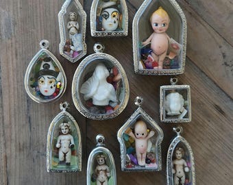 May include: A collection of 10 silver framed pendants with glass windows. Each pendant contains a small porcelain doll or figurine, some with colorful gems and beads. The pendants are arranged on a wooden surface.