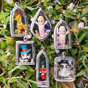 May include: Six silver framed pendants with glass fronts, each containing a different miniature figurine. The figurines include a giraffe, a baby, a clown, a soldier, and two other figures. The pendants are arranged on a bed of green grass and pink flowers.