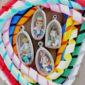 Puede incluir: Cuatro colgantes plateados con figuras de muñecas en miniatura en su interior, rodeadas de piedras de colores. Los colgantes se colocan sobre un fondo de cintas en espiral de colores brillantes: rojo, blanco, azul, rosa, amarillo y verde.