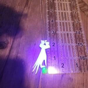 May include: A silver chain necklace with a white pendant shaped like a hand holding a green gemstone. The pendant is approximately 2 inches tall.