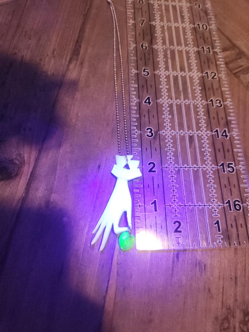 May include: A silver chain necklace with a white pendant shaped like a hand holding a green gemstone. The pendant is approximately 2 inches tall.