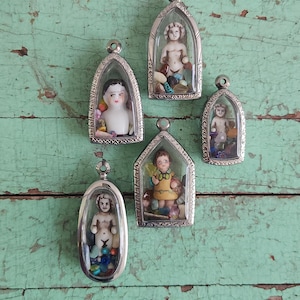 Puede incluir: Una colección de colgantes plateados antiguos, cada uno con una pequeña figura. Las figuras incluyen una muñeca de porcelana y figuras de querubines, rodeadas de cuentas de colores y objetos pequeños. Los colgantes se exhiben sobre una superficie de madera pintada de color turquesa desgastado.