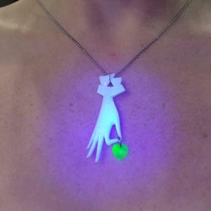 May include: A white ceramic necklace with a hand holding a green heart pendant. The necklace is on a person's chest and the pendant is glowing green under a black light.