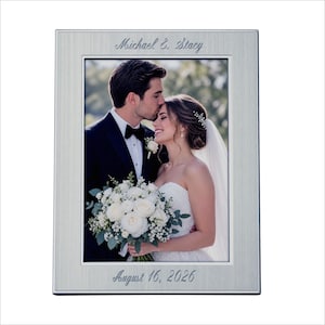 May include: A rectangular brushed silver picture frame featuring engraved text at the top reading Michael & Stacy and at the bottom reading August 16, 2026. The frame contains a photograph of a couple in wedding attire embracing outdoors.