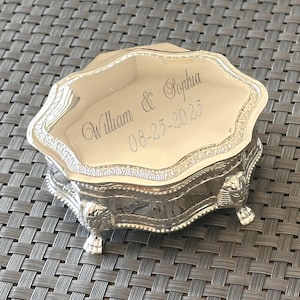 May include: A silver jewellery box with intricate detailing and a cream interior. The box is engraved with the names "William & Sophia" and the date "08-23-2025". The box has decorative feet.