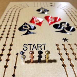 May include: A wooden game board with a "START" label and four pegs in different colors: silver, copper, black, and gold. The board has a design of playing cards with the number 5 of spades.