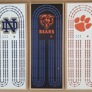 May include: Three wooden signs with different college logos and team names. The first sign has a blue and gold Notre Dame Fighting Irish logo with the text "ND". The second sign has a black background with a blue and white chain design and the text "BEARS". The third sign has a white background with an orange paw print and the text "CLEMSON".