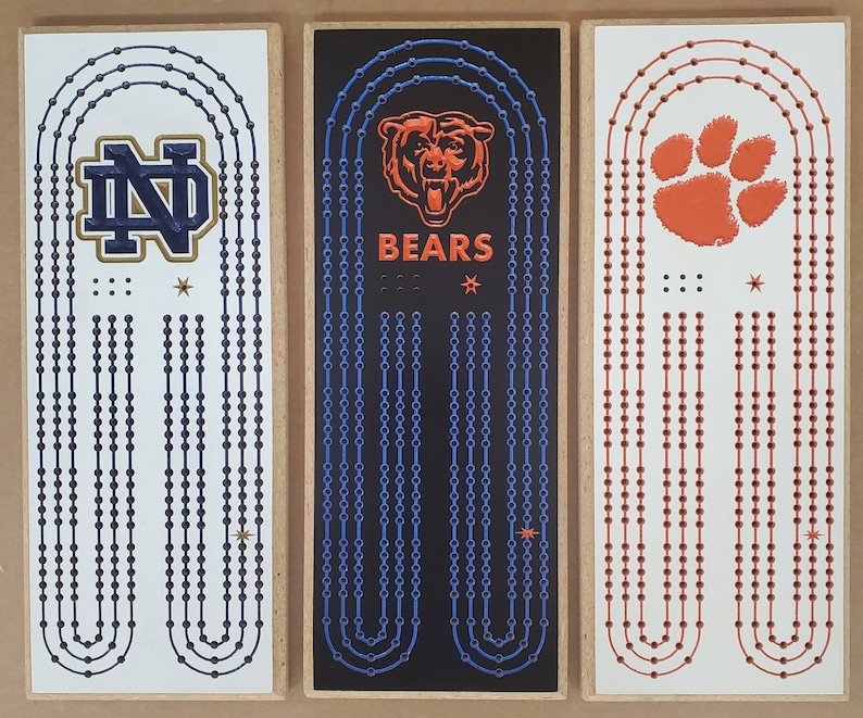 May include: Three wooden signs with different college logos and team names. The first sign has a blue and gold Notre Dame Fighting Irish logo with the text "ND". The second sign has a black background with a blue and white chain design and the text "BEARS". The third sign has a white background with an orange paw print and the text "CLEMSON".