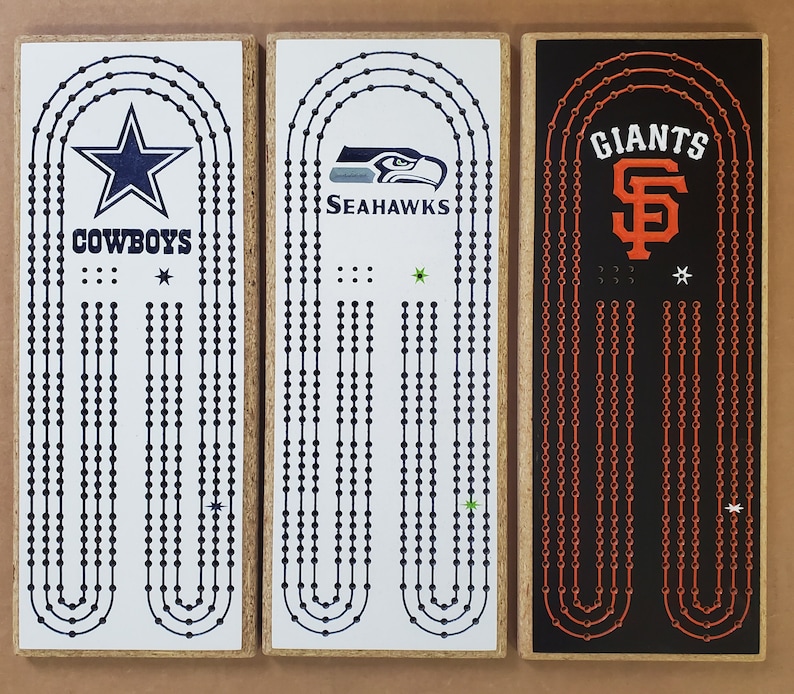 May include: Three wooden game boards with different NFL and MLB team logos. The boards are white, black, and brown with black, blue, green, orange, and red details. The logos are for the Dallas Cowboys, Seattle Seahawks, and San Francisco Giants.