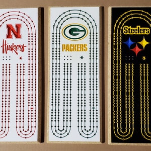 May include: Three wooden game boards with different team logos. The first board is white with red dots and the logo for the Nebraska Huskers. The second board is white with green dots and the logo for the Green Bay Packers. The third board is black with gold dots and the logo for the Pittsburgh Steelers.