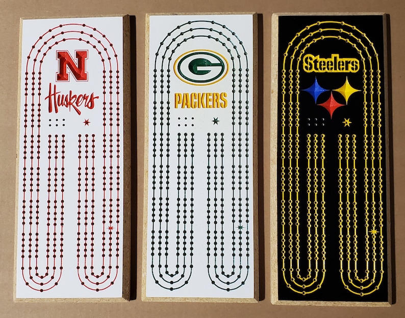 May include: Three wooden game boards with different team logos. The first board is white with red dots and the logo for the Nebraska Huskers. The second board is white with green dots and the logo for the Green Bay Packers. The third board is black with gold dots and the logo for the Pittsburgh Steelers.