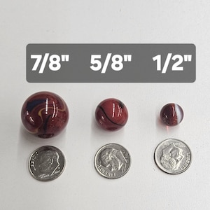 May include: Three round glass marbles in shades of deep red with dark swirls, arranged by size next to three US dimes for scale. Text labels above the marbles indicate diameters of 7/8 inch, 5/8 inch, and 1/2 inch respectively.