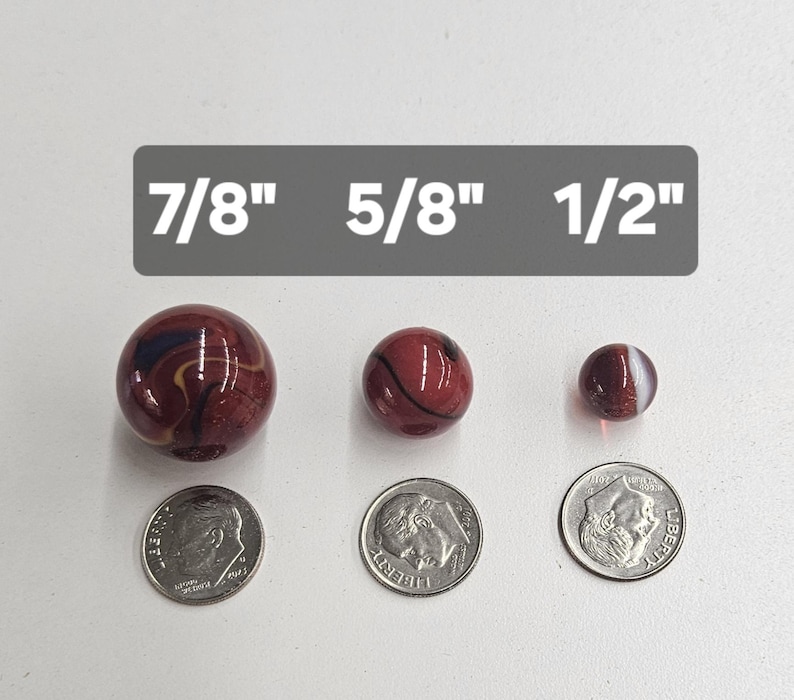 May include: Three round glass marbles in shades of deep red with dark swirls, arranged by size next to three US dimes for scale. Text labels above the marbles indicate diameters of 7/8 inch, 5/8 inch, and 1/2 inch respectively.