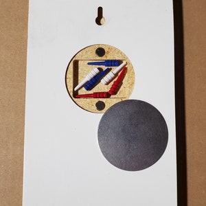 May include: A white wooden board with a round cutout revealing a set of red, white, and blue wooden pegs. A round metal disc is attached to the board.