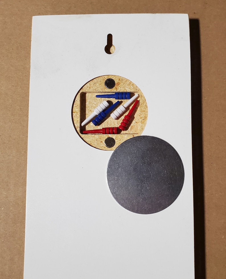 May include: A white wooden board with a round cutout revealing a set of red, white, and blue wooden pegs. A round metal disc is attached to the board.