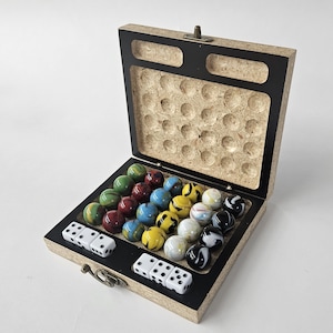 Pode incluir: Uma caixa de jogo de madeira com interior preto, contendo bolas de gude coloridas e dados brancos. As bolas de gude são de várias cores, incluindo verde, vermelho, azul, amarelo e preto e branco. A caixa está aberta, revelando as peças do jogo.
