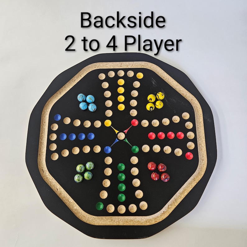 May include: An octagonal black game board with a light-colored wooden border, featuring a cross-shaped path of indentations for marbles. The text "Backside 2 to 4 Player" is printed above. Marbles in blue, yellow, red, and green are placed on the board.
