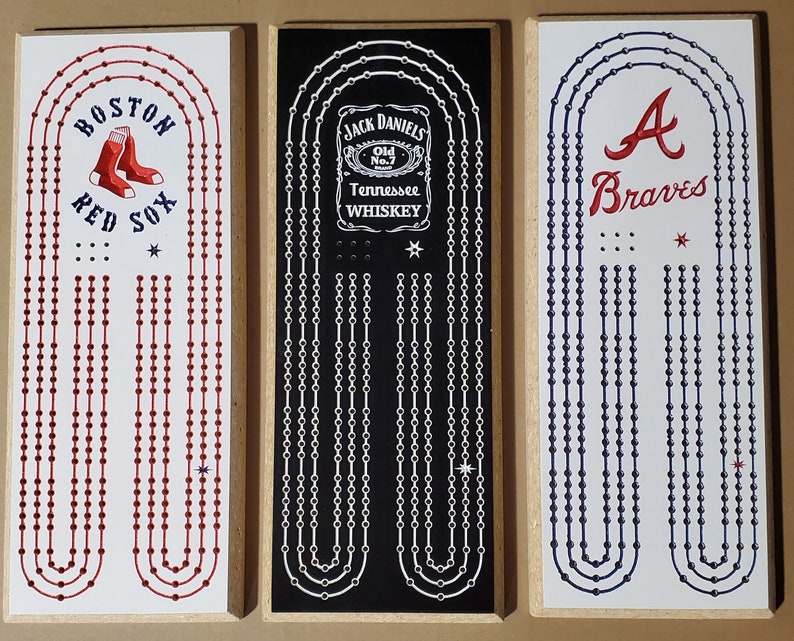 May include: Three wooden game boards with different team logos and names. The first board has a red and white Boston Red Sox logo. The second board has a black and white Jack Daniels logo. The third board has a blue and white Atlanta Braves logo.