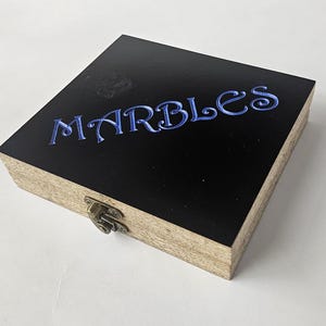 May include: A square wooden box with a black lid featuring the word MARBLES engraved in blue lettering. The sides of the box show a natural light brown wood grain texture, and a small metallic latch is attached to the front for closure.