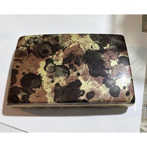 Peut inclure: Une boîte rectangulaire présentant un motif de formes organiques abstraites dans des tons de brun foncé, de noir et d'or métallique. Le couvercle a une finition lisse et brillante avec un design continu qui recouvre toute la surface supérieure.