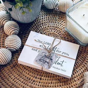 May include: A stack of white cards tied with twine, featuring the text "The LORD will fight for you, and you shall be still." A decorative tag is attached. The cards are on a woven tray, with a candle and decorative wooden beads.