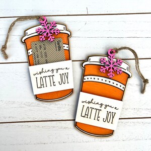 Puede incluir: Dos adornos de taza de caf&eacute; naranja y blanca con un copo de nieve rosa en la parte superior. Cada adorno tiene una etiqueta blanca que dice "wishing you a LATTE JOY".