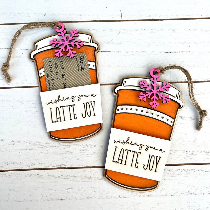 Puede incluir: Dos adornos de taza de caf&eacute; naranja y blanca con un copo de nieve rosa en la parte superior. Cada adorno tiene una etiqueta blanca que dice "wishing you a LATTE JOY".