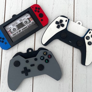 May include: Three wooden ornaments shaped like video game controllers. The first is a blue and red Nintendo Switch, the second is a white and black Playstation 5 controller, and the third is a gray Xbox controller. Each ornament has a hole at the top for hanging.