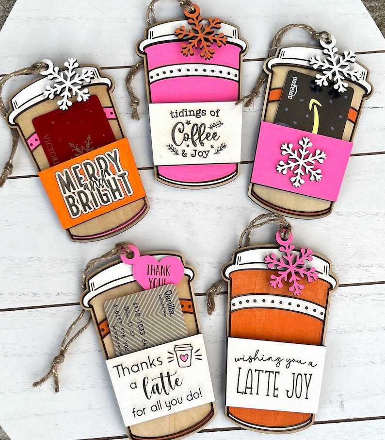 Puede incluir: Cuatro adornos de madera en forma de taza de caf&eacute; con tarjetas de regalo y mensajes festivos. Los adornos est&aacute;n decorados con copos de nieve, corazones y las palabras "Merry and Bright", "Tidings of Coffee & Joy", "Thanks a Latte for all you do!" y "Wishing you a Latte Joy".