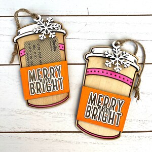 Puede incluir: Dos adornos de madera en forma de taza de caf&eacute; con un dise&ntilde;o a rayas rosa y blanco. Los adornos tienen un porta tarjetas de regalo y un mensaje "Merry and Bright" en la manga naranja.