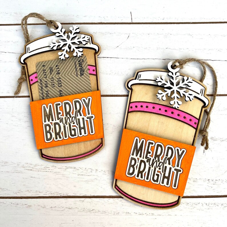 Puede incluir: Dos adornos de madera en forma de taza de caf&eacute; con un dise&ntilde;o a rayas rosa y blanco. Los adornos tienen un porta tarjetas de regalo y un mensaje "Merry and Bright" en la manga naranja.