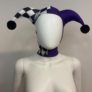 Peut inclure: Un chapeau de bouffon avec un motif à carreaux noir et blanc d'un côté et violet uni de l'autre. Le chapeau a trois pointes, chacune avec un pompon noir. Le tour de cou est également à carreaux et violet.