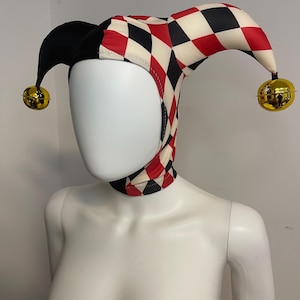 Peut inclure: Chapeau de bouffon avec un motif de losanges noirs, rouges et crème. Le chapeau a deux pointes noires et des cloches dorées aux extrémités. Le chapeau est porté par un mannequin.