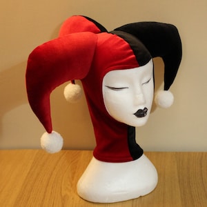 Peut inclure: Un chapeau de jester en velours rouge et noir avec des pompons blancs aux extrémités des cornes. Le chapeau est sur une tête de mannequin blanche.