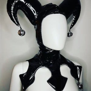 Puede incluir: Conjunto de capucha y cuello de bufón de vinilo negro. La capucha presenta puntas altas y curvas con campanas plateadas en los extremos. El cuello tiene un diseño en forma de diamante con campanas plateadas. El conjunto se muestra en un maniquí.