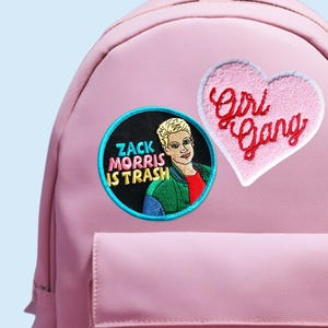 May include: A pink backpack with a front pocket and a top handle. The backpack features three embroidered patches: a heart-shaped patch with "Girl Gang" text, a round patch with a portrait and "Zack Morris Is Trash" text, and a patch that says "professional dog petter."