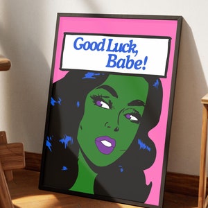 May include: A framed print with a pink background and a black-haired woman with green skin. The woman is looking to the right and has purple lips. The text "Good Luck, Babe!" is in a white speech bubble above her head.