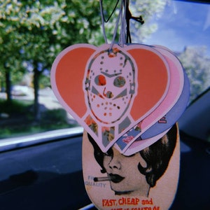 May include: A set of three car air fresheners hanging from a rearview mirror. The first air freshener is a pink heart with a white hockey mask design. The second air freshener is a pink and white heart with a floral pattern. The third air freshener is a light pink and white oval with a woman's face and the text "Fast, Cheap and Out of Control".