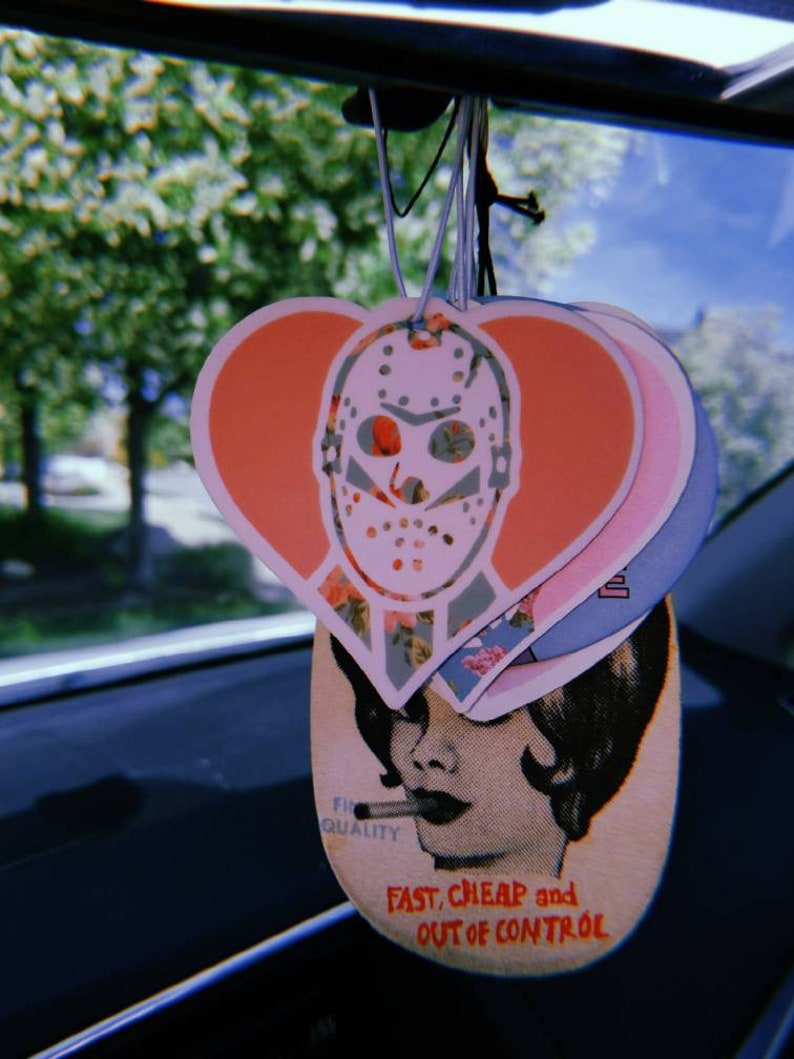 May include: A set of three car air fresheners hanging from a rearview mirror. The first air freshener is a pink heart with a white hockey mask design. The second air freshener is a pink and white heart with a floral pattern. The third air freshener is a light pink and white oval with a woman's face and the text "Fast, Cheap and Out of Control".