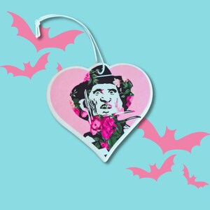 May include: A pink heart-shaped car air freshener with a floral design featuring a character from a horror movie. The character is wearing a brown hat and has a knife in their hand.