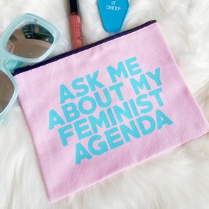 May include: A pink canvas makeup bag with a blue inscription that reads "Ask Me About My Feminist Agenda".