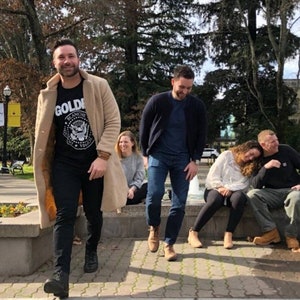 May include: A group of four people standing in front of a fountain. The person at the front is wearing a tan coat over a black t-shirt with the text "GOLD" and a graphic of an eagle. The person behind them is wearing a blue shirt and jeans. The two people at the back are sitting on a stone wall.