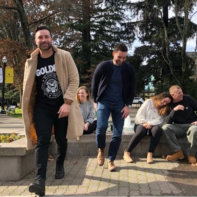 May include: A group of four people standing in front of a fountain. The person at the front is wearing a tan coat over a black t-shirt with the text "GOLD" and a graphic of an eagle. The person behind them is wearing a blue shirt and jeans. The two people at the back are sitting on a stone wall.