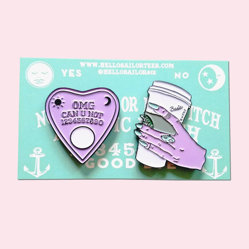 Op de afbeelding: Twee emaille pinnen. De eerste is een paarse hartvormige Ouija-plank met de tekst "OMG CAN U NOT 1234567890". De tweede is een paarse hand die een witte koffiebeker vasthoudt met de tekst "Basic Bucks".