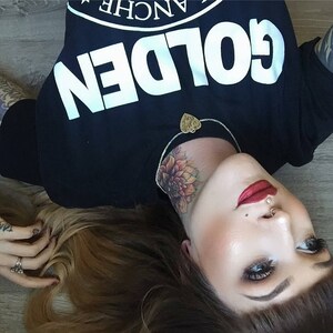 May include: A woman with long brown hair is lying on her back. She is wearing a black t-shirt with the word "GOLDEN" printed in white on the front. She has a tattoo of a flower on her neck and is wearing a necklace with a heart-shaped pendant. Her lips are painted a bright red.