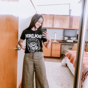 May include: A woman wearing a black t-shirt with a white graphic of the presidential seal and the word "GOLDEN" printed backwards. She is also wearing plaid wide-leg trousers.