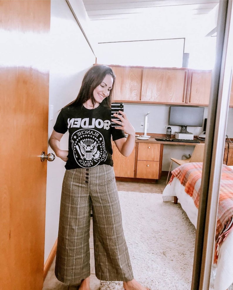 May include: A woman wearing a black t-shirt with a white graphic of the presidential seal and the word "GOLDEN" printed backwards. She is also wearing plaid wide-leg trousers.