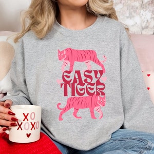 K&ouml;nnte beinhalten: Ein graues Sweatshirt mit einem rosafarbenen Tiger-Motiv und dem Text "EASY TIGER" in einer Retro-Schrift. Das Sweatshirt wird von einer Person getragen, die eine wei&szlig;e Tasse mit roten Herzen und dem Text "XOXO" h&auml;lt.