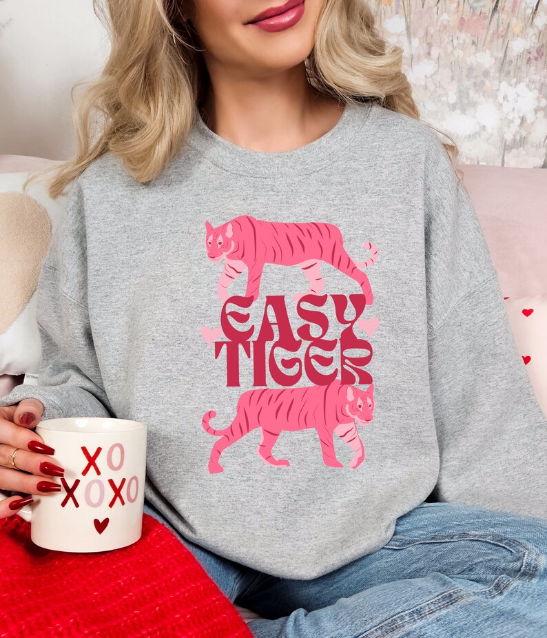 K&ouml;nnte beinhalten: Ein graues Sweatshirt mit einem rosafarbenen Tiger-Motiv und dem Text "EASY TIGER" in einer Retro-Schrift. Das Sweatshirt wird von einer Person getragen, die eine wei&szlig;e Tasse mit roten Herzen und dem Text "XOXO" h&auml;lt.