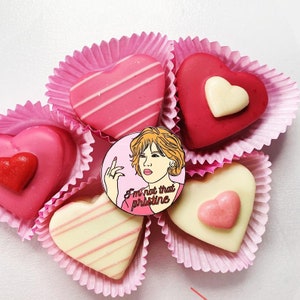 Puede incluir: Un pin de esmalte rosa y blanco con una imagen de dibujos animados de una mujer con cabello rojo y el texto "I'm not that pristine". El pin est&aacute; rodeado de chocolates en forma de coraz&oacute;n rosa en vasos de papel rosa.